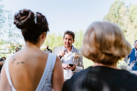 Hombre con traje rayado habla con un micrófono frente a una mujer con vestido blanco y una mujer mayor vestida de negro.