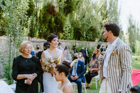 Hombre con traje rayado habla con un micrófono junto a una mujer con vestido blanco y una mujer mayor vestida de negro.