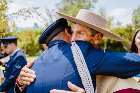 Hombre con uniforme azul abraza a un hombre con sombrero claro al aire libre, con otras personas alrededor.