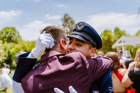 Hombre con uniforme azul abraza a un hombre con traje color burdeos al aire libre.