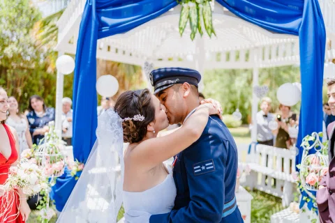 Hombre con uniforme azul besa a una mujer con vestido blanco frente a una estructura decorada.