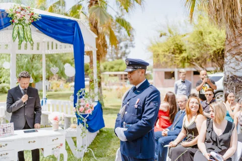 Hombre con uniforme azul y gorra permanece de pie junto a una estructura decorada en azul y blanco, con personas sentadas detrás.