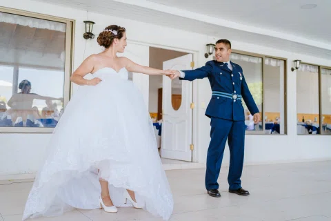 Hombre con uniforme azul y mujer con vestido blanco bailan tomados de una mano en un espacio interior.