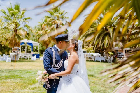 Hombre con uniforme azul y mujer con vestido blanco se besan en un jardín, junto a palmeras.