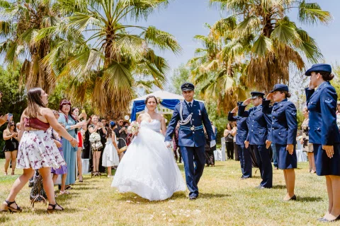 Hombre con uniforme azul y mujer con vestido blanco caminan por el jardín entre varias personas uniformadas.