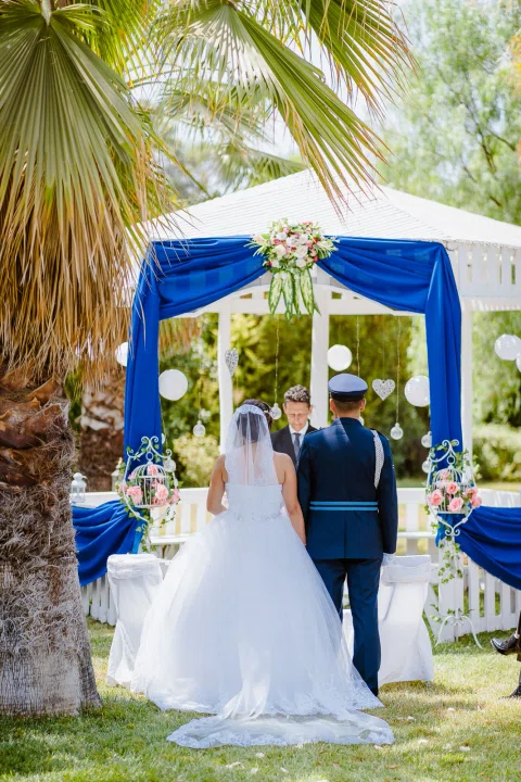 Hombre con uniforme azul y mujer con vestido blanco, vistos de espaldas, permanecen frente a un hombre con traje oscuro bajo una estructura decorada.