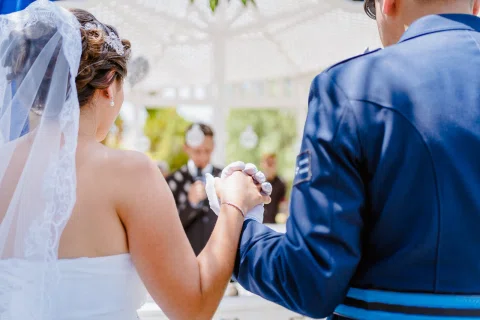 Hombre con uniforme azul y mujer con vestido blanco son vistos de espaldas con las manos unidas frente a ellos.