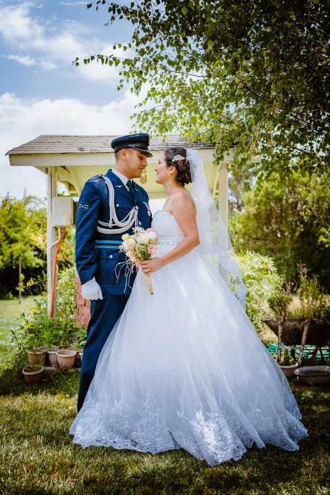 Hombre con uniforme azul y mujer con vestido blanco y ramo posan juntos en un jardín.