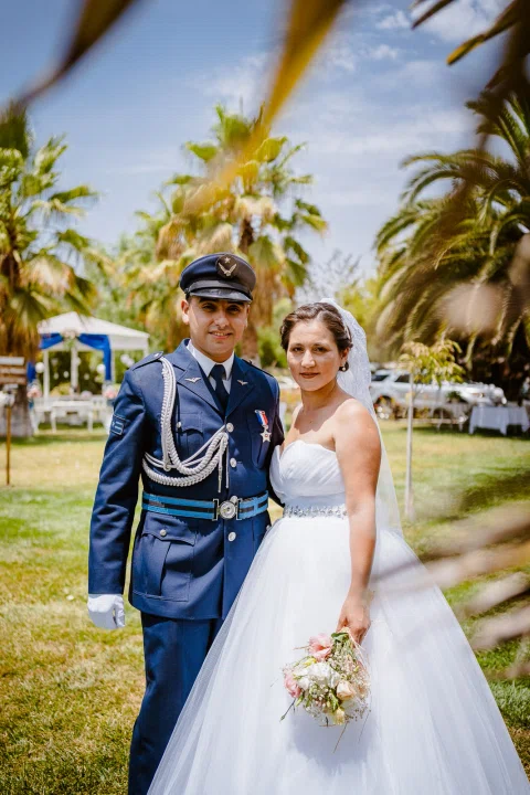 Hombre con uniforme azul y mujer con vestido blanco sosteniendo un ramo posan juntos al aire libre.
