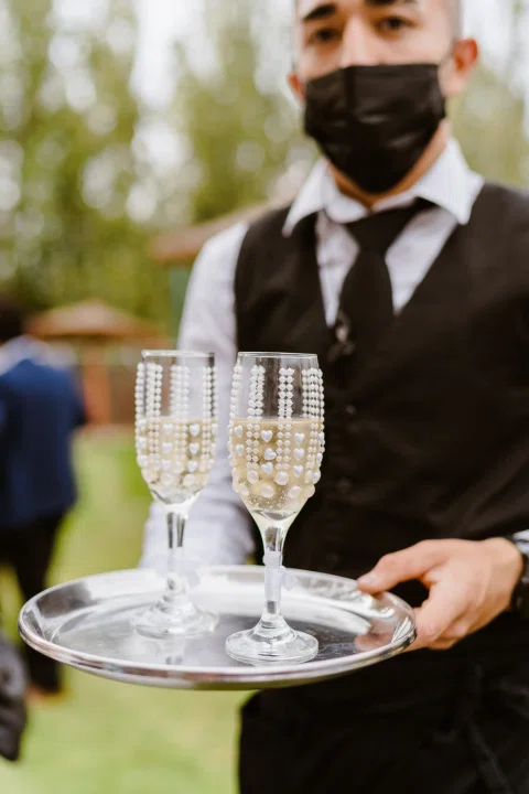 Hombre con uniforme negro y mascarilla sosteniendo una bandeja con dos copas.