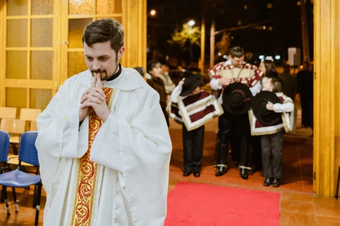 Hombre con vestimenta litúrgica mantiene las manos juntas frente al rostro dentro de una iglesia, con otras personas detrás.