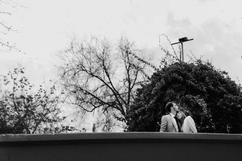 Hombre y mujer aparecen a distancia detrás de un muro bajo los árboles, en una fotografía en blanco y negro.