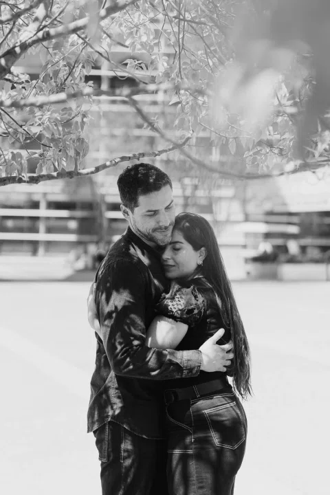 Hombre y mujer se abrazan en una fotografía en blanco y negro con vegetación desenfocada.