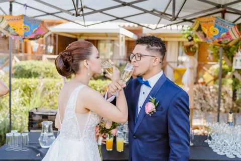 Mujer con vestido blanco y hombre con traje azul beben de copas con los brazos entrelazados.