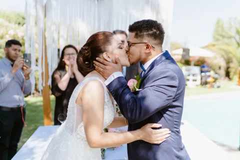 Mujer con vestido blanco y hombre con traje azul se besan frente a una mesa, con una piscina al fondo.