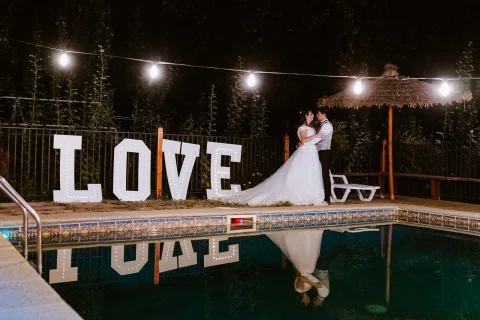 Hombre y mujer se besan junto a las letras LOVE iluminadas al borde de una piscina durante la noche.