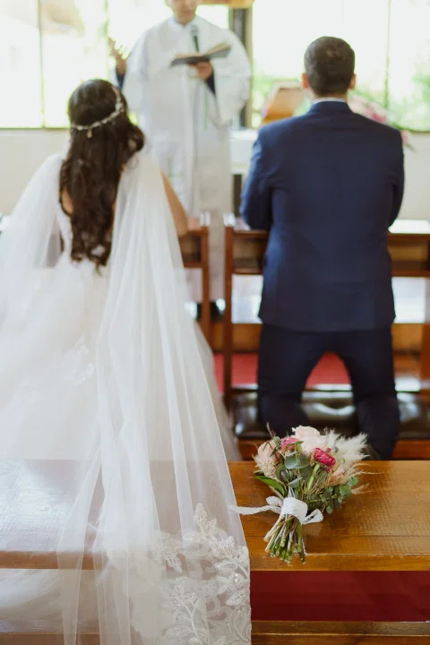 Hombre con traje azul y mujer con vestido blanco, vistos de espaldas, permanecen frente a un hombre que sostiene un libro dentro de la iglesia.