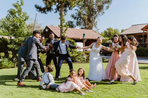 Mujer con vestido blanco y hombre con traje azul estiran los brazos mientras varias personas los sujetan desde lados opuestos; tres niños están sentados sobre el césped en primer plano.