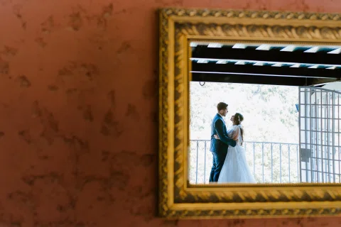 Hombre con traje azul y mujer con vestido blanco aparecen reflejados en un espejo de marco dorado junto a un balcón.