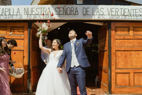 Hombre con traje azul y mujer con vestido blanco salen por la puerta de la capilla con los brazos levantados.