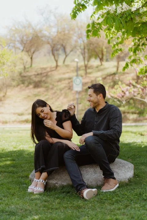 Hombre y mujer están sentados sobre una piedra; ella ríe mientras él la mira.