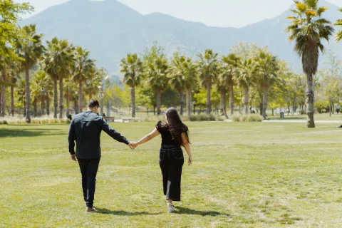 Hombre y mujer caminan tomados de la mano por un parque con palmeras y montañas al fondo.