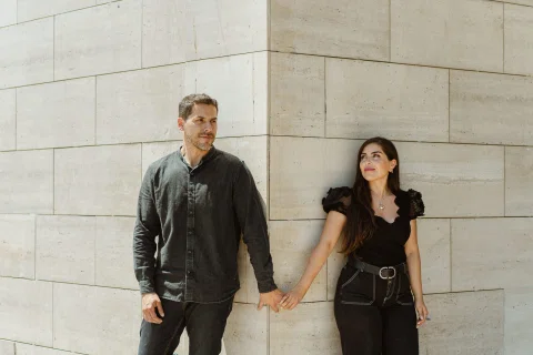 Hombre y mujer permanecen tomados de la mano junto a un muro de piedra clara.
