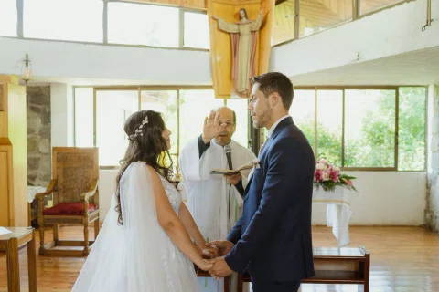 Hombre con traje azul y mujer con vestido blanco se toman de las manos frente a un hombre con vestimenta clara que sostiene un libro.