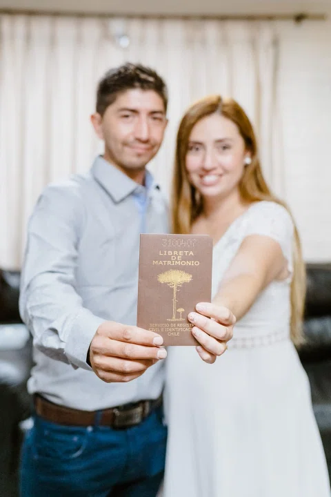 Libreta de Matrimonio sostenida en primer plano, con un hombre de camisa celeste y una mujer de vestido blanco desenfocados al fondo.