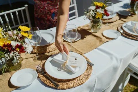 Mano ajusta una servilleta sobre un plato en una mesa exterior con flores y copas.