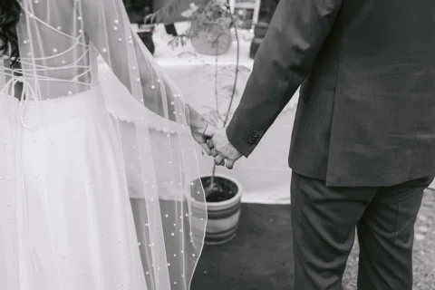 Detalle en blanco y negro de las manos entrelazadas de una mujer con vestido blanco y un hombre con traje oscuro.