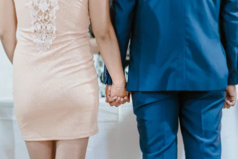 Detalle de dos manos entrelazadas junto a un vestido rosado y un traje azul.