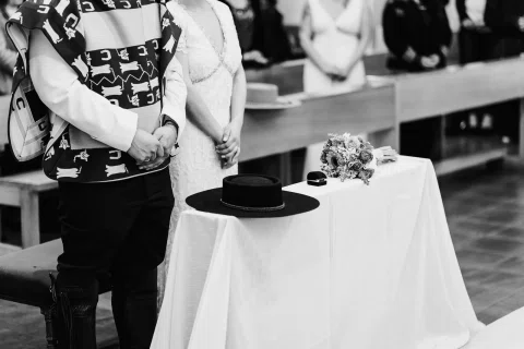 Manos de un hombre con poncho y una mujer con vestido blanco se encuentran junto a una mesa con sombrero, ramo y una caja pequeña, en una fotografía en blanco y negro.