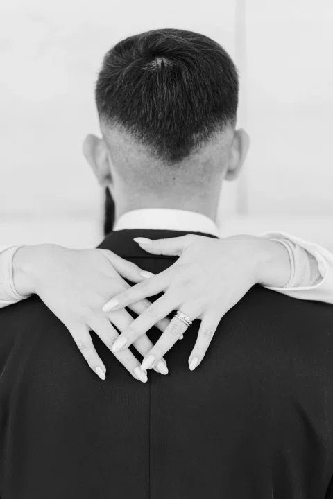 Detalle en blanco y negro de las manos de una mujer apoyadas y cruzadas sobre la espalda de un hombre.