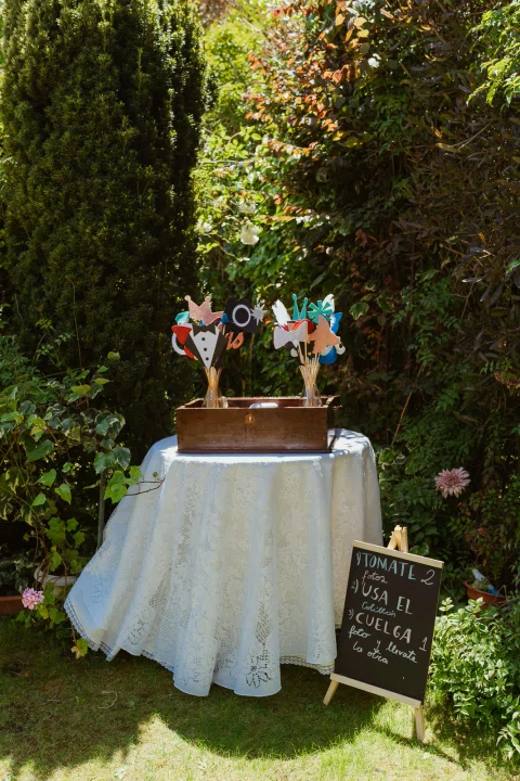 Mesa redonda con accesorios de utilería para fotografías y un pequeño cartel de instrucciones en el jardín.