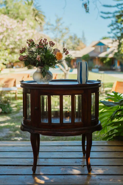 Mesa redonda de madera con un ramo de flores y una jarra metálica sobre una terraza.