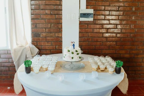 Mesa con torta, dulces y decoración blanca frente a un muro de ladrillo.