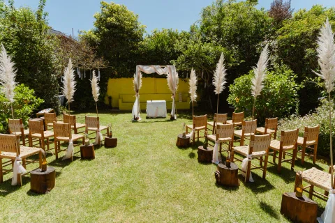 Montaje de ceremonia al aire libre con sillas de madera y un arco blanco al fondo.