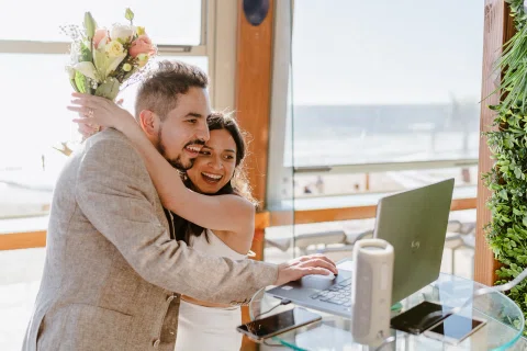 Mujer abraza a un hombre junto a un computador y un ventanal con vista al mar.