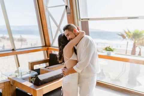Mujer abraza a un hombre de pie junto a una ventana con vista al mar.