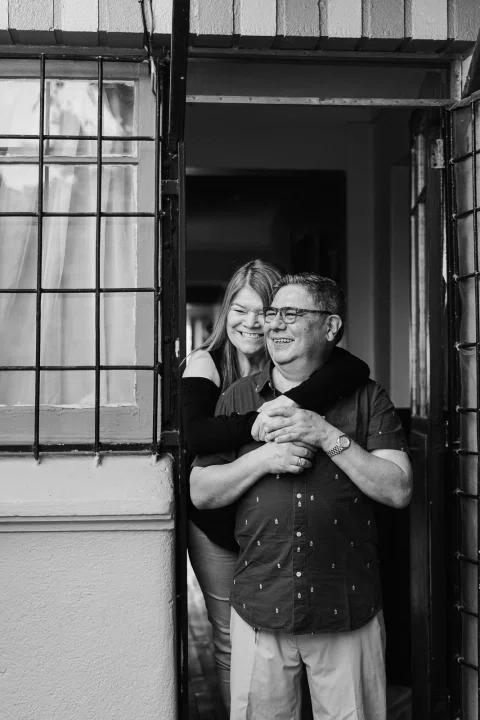 Mujer abraza desde atrás a un hombre en una puerta, en una fotografía en blanco y negro.