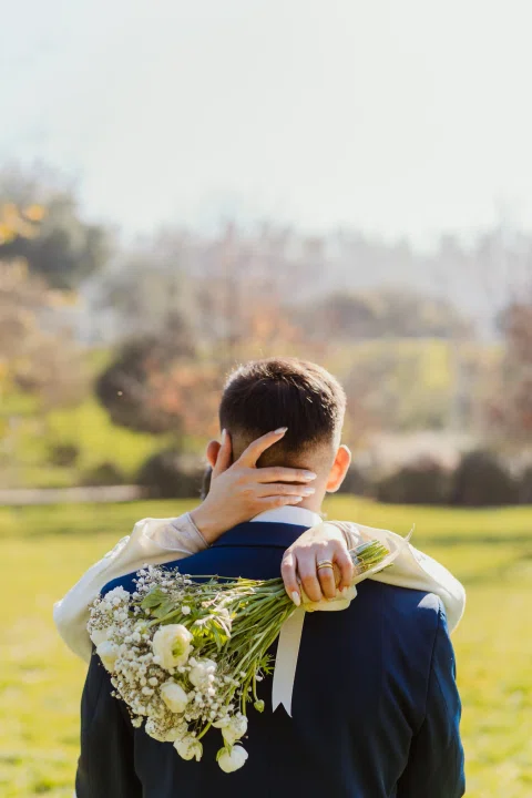 Mujer con traje claro abraza por el cuello a un hombre con traje azul mientras sostiene un ramo de flores blancas.