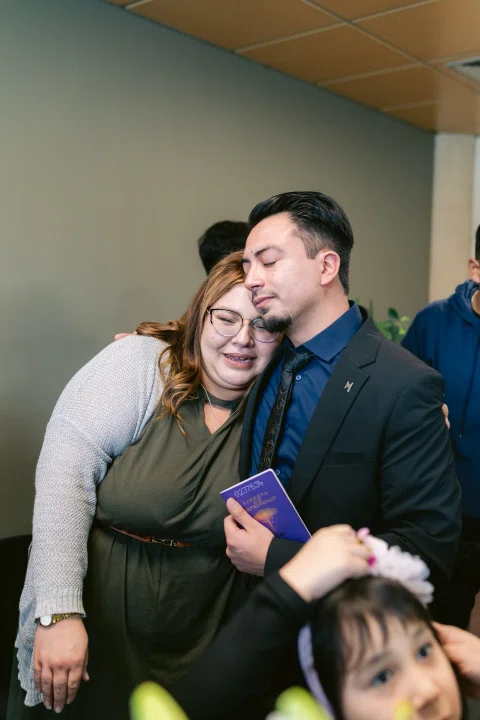Mujer abraza a un hombre de traje negro que sostiene un cuadernillo morado, con un niño visible en primer plano.
