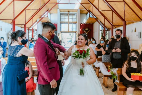 Mujer con vestido azul ajusta la chaqueta de un hombre con traje burdeos mientras una mujer con vestido blanco sostiene un ramo.