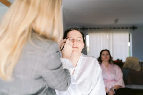 Mujer aplica maquillaje a otra mujer sentada con una bata blanca.