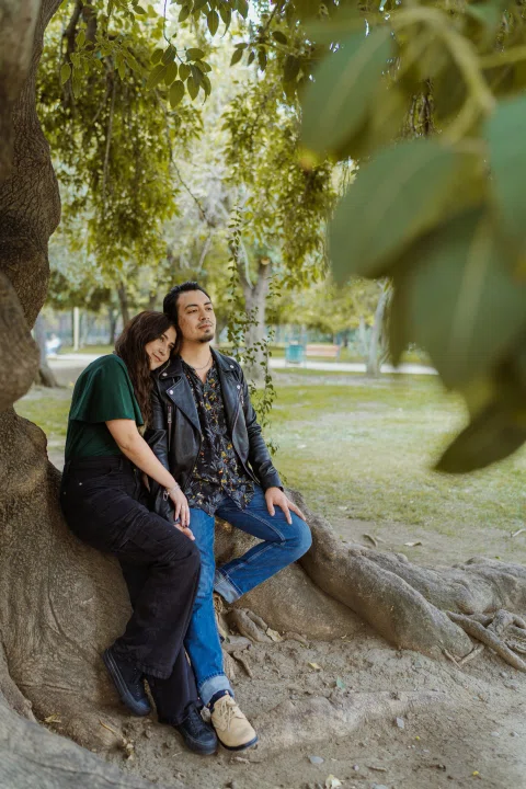 Mujer apoya la cabeza junto a un hombre mientras ambos están sentados sobre las raíces de un árbol.