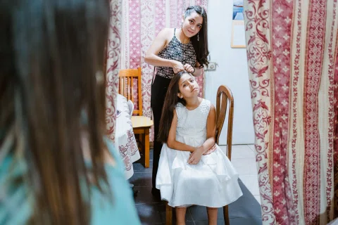Mujer con vestido oscuro arregla el cabello de una niña con vestido blanco sentada en una silla.