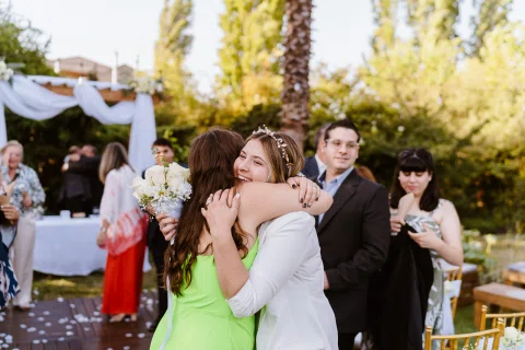 Mujer vestida de blanco abraza a una mujer con vestido verde en un jardín.