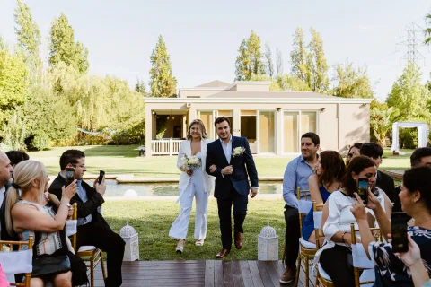 Mujer vestida de blanco y hombre con traje azul caminan juntos por un pasillo entre personas sentadas al aire libre.