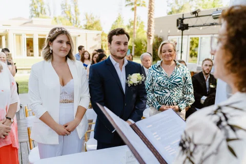 Mujer vestida de blanco y hombre con traje azul permanecen de pie frente a una persona en un espacio exterior.